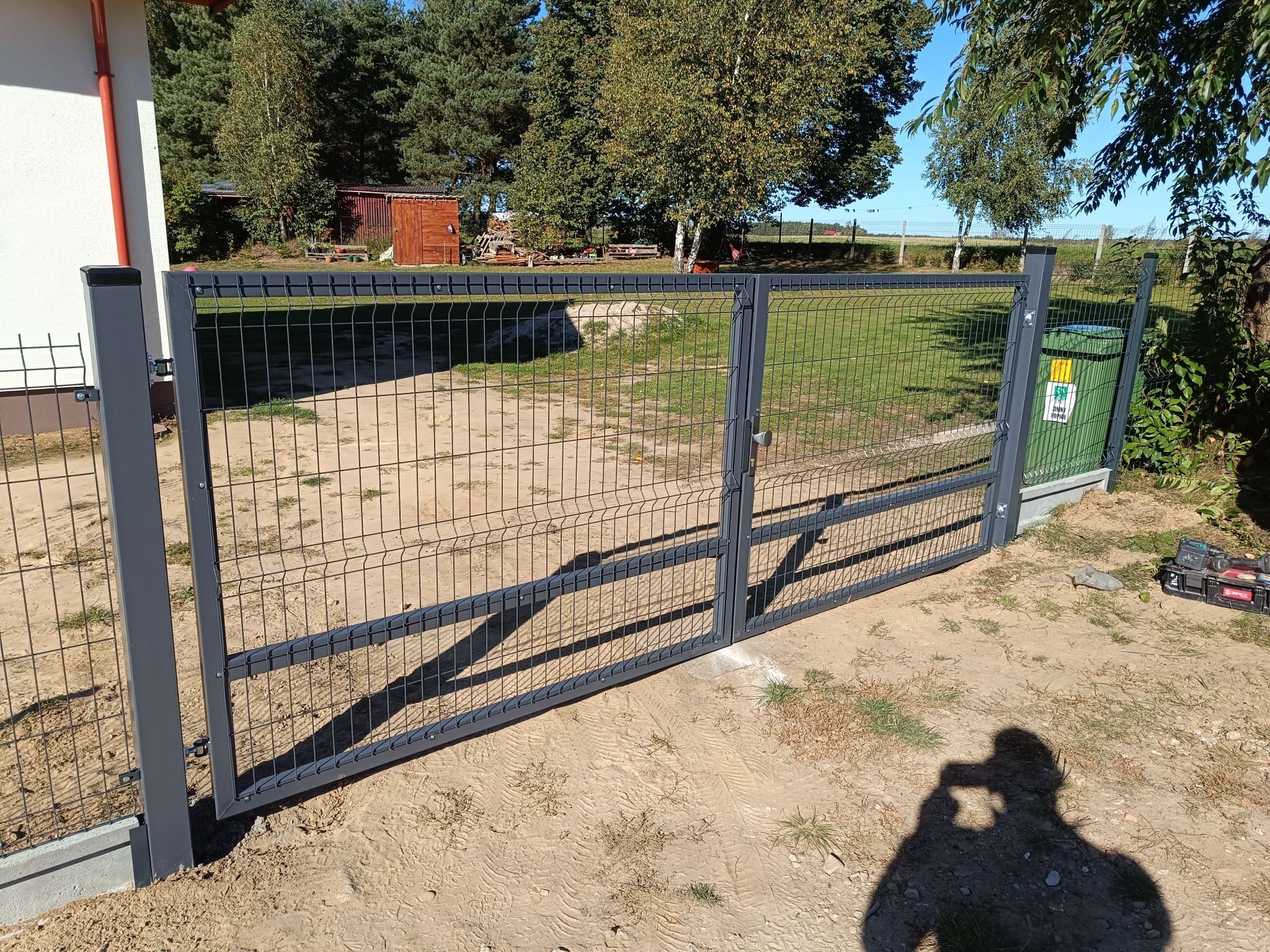Szara, metalowa brama panelowa, dwuskrzydłowa, z widocznym słupkiem i narzędziami montażowymi obok. Tło: trawa, drzewa, budynek. Cień osoby robiącej zdjęcie.