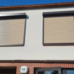 Elewacja domu z dwoma oknami z roletami zewnętrznymi w kolorze beżowym, jedna roleta częściowo podniesiona, widoczny numer domu 22 i fragment balkonu.