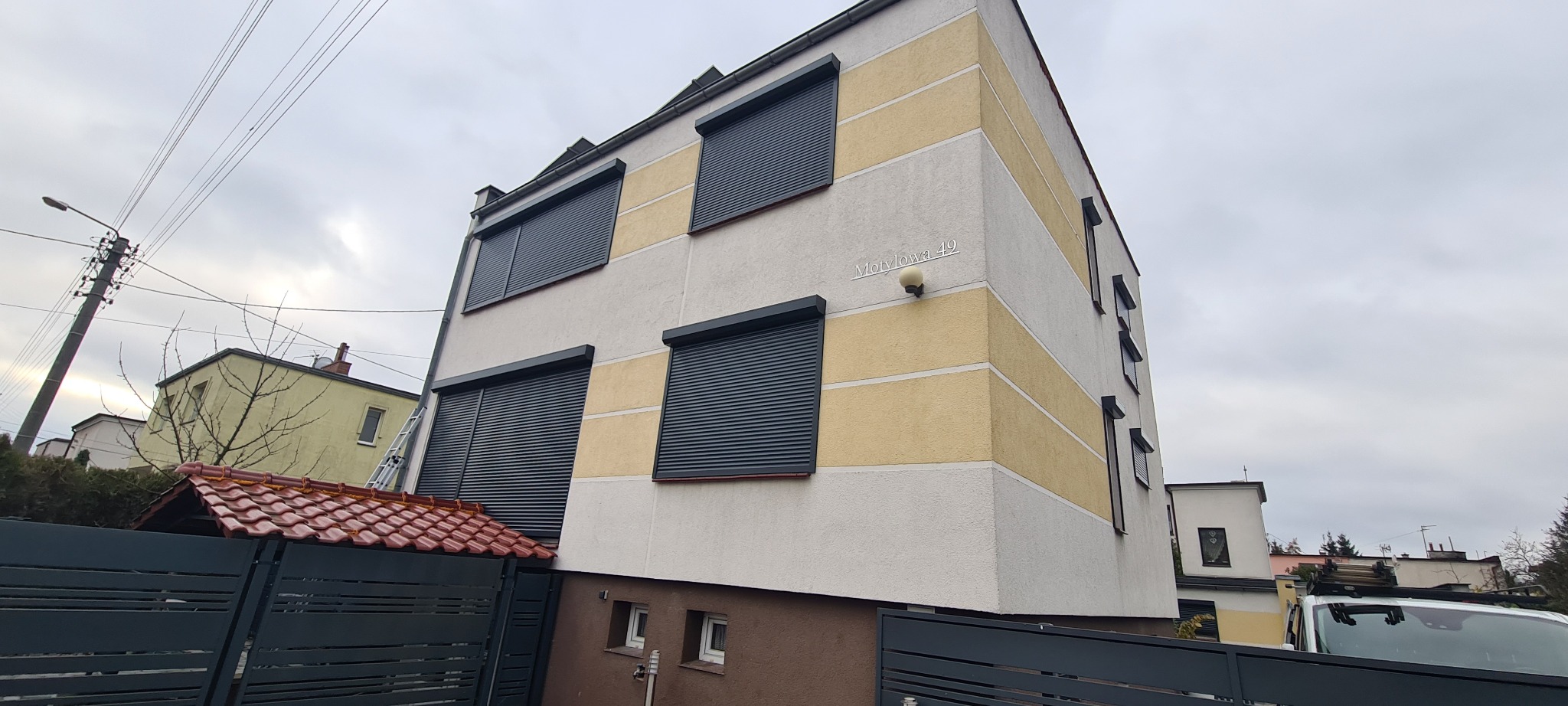 Dwupiętrowy budynek z zamontowanymi, zamkniętymi, grafitowymi osłonami okiennymi, widocznymi z zewnątrz, z drabiny opartą o ścianę, adres 'Motylowa 49'.
