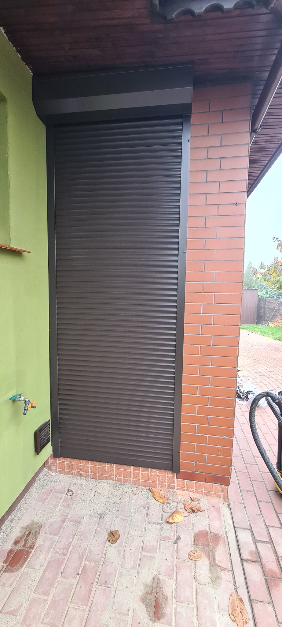 Zamknięta, brązowa roleta zewnętrzna zamontowana na ścianie budynku z cegły i zieloną ścianą, widoczne liście na chodniku.