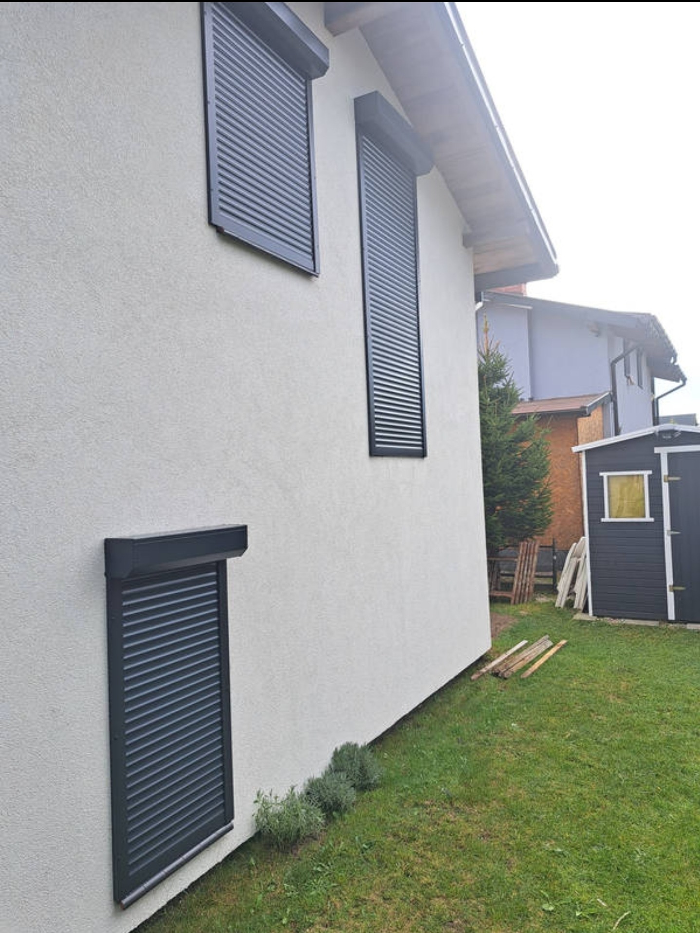 Elewacja domu z trzema oknami zabezpieczonymi grafitowymi roletami zewnętrznymi w pozycji zamkniętej, widok z boku na trawnik i domek narzędziowy.