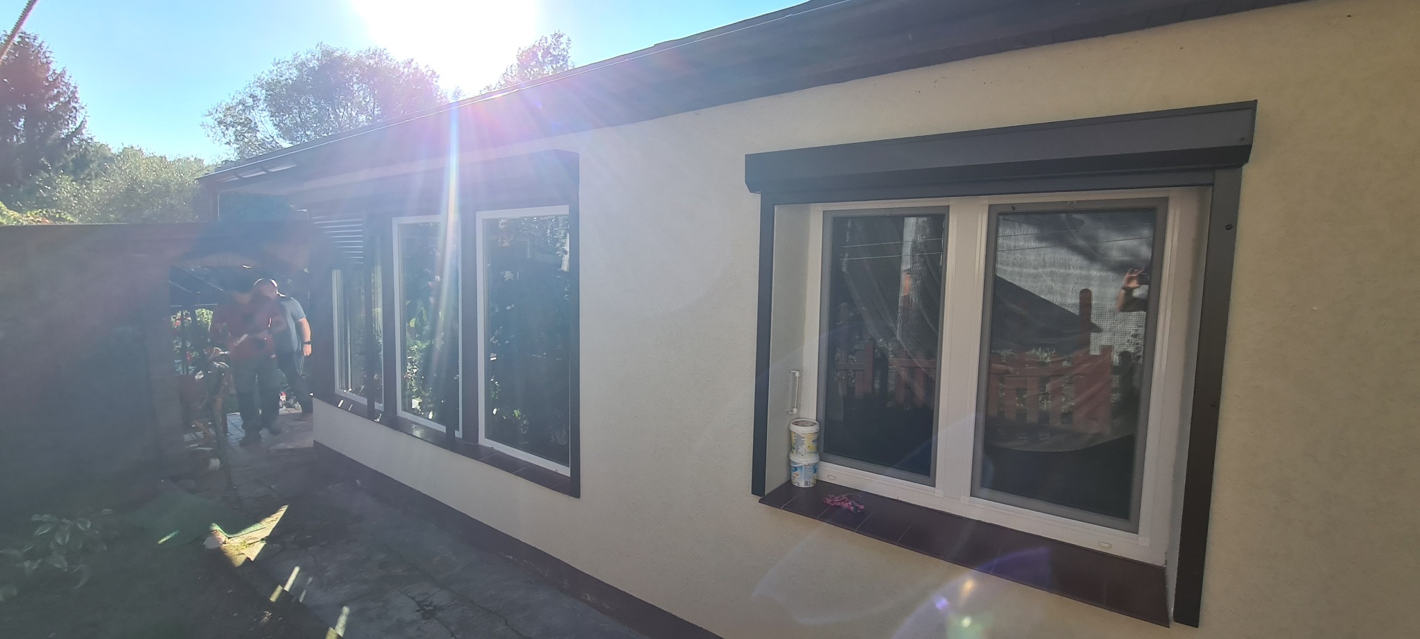 Zamknięta, szara roleta zewnętrzna na oknie budynku, z widocznymi dwoma puszkami farby na parapecie i odbiciem fotografa w szybie.