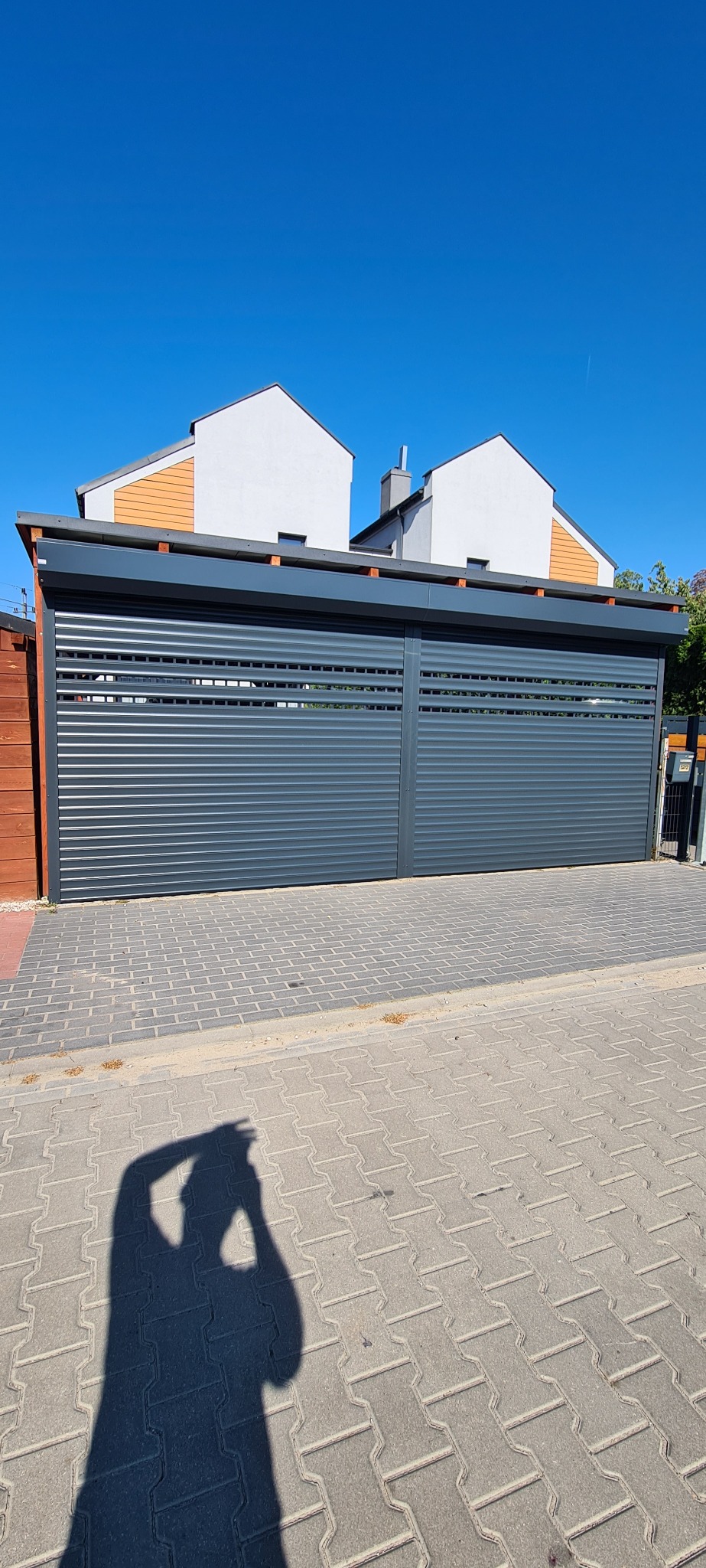 Szara, pozioma brama garażowa z lamelami, widok z zewnątrz, na pierwszym planie cień osoby robiącej zdjęcie, w tle dwa budynki mieszkalne pod błękitnym niebem.