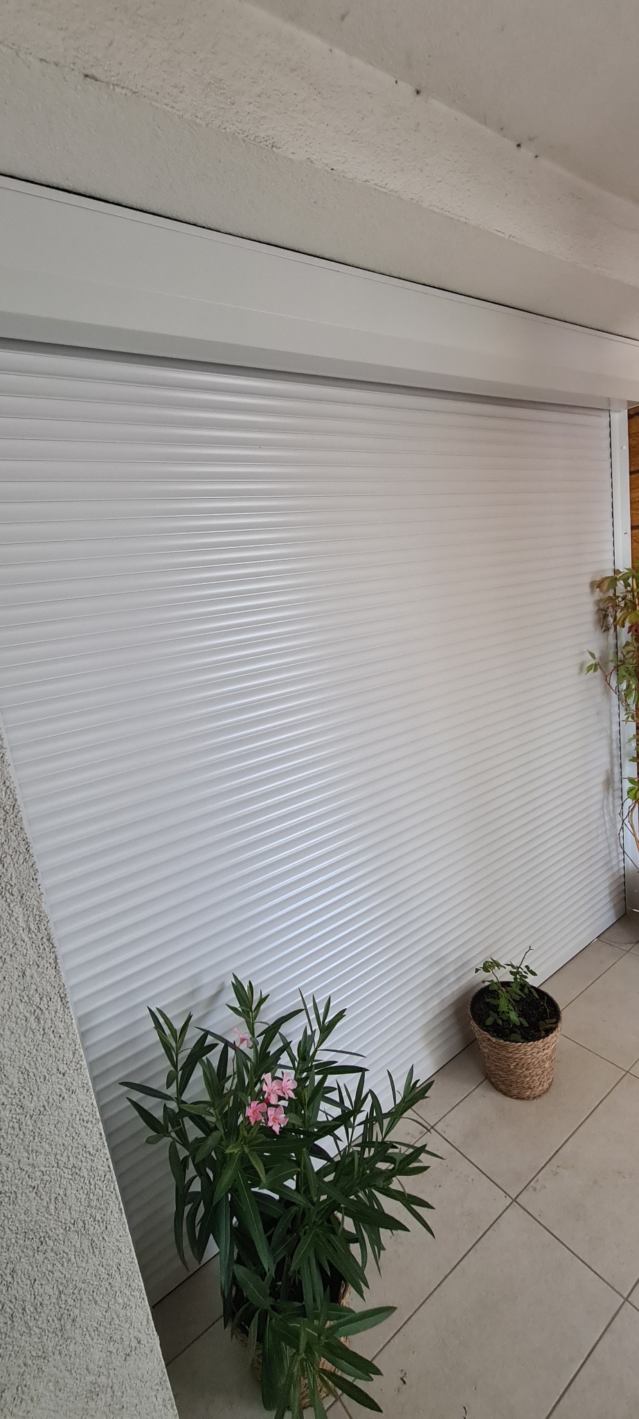 Biała, pozioma osłona okienna z delikatnymi żłobieniami, częściowo opuszczona, z doniczkami z roślinami po bokach, na balkonie z jasnymi płytkami.