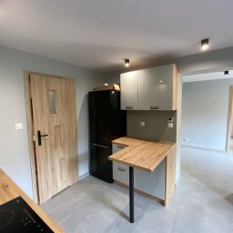 NAZBUD NAZARII HRYTSYNA - Jasna kuchnia z drewnianym blatem i szarymi szafkami, czarna lodówka, szara podłoga, drewniane drzwi z przeszkleniem. Nowoczesne oświetlenie punktowe.