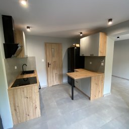 NAZBUD NAZARII HRYTSYNA - Nowoczesna kuchnia z drewnianymi akcentami: zabudowa kuchenna z blatem, czarna płyta indukcyjna i okap, szare płytki podłogowe, minimalistyczne oświetlenie.