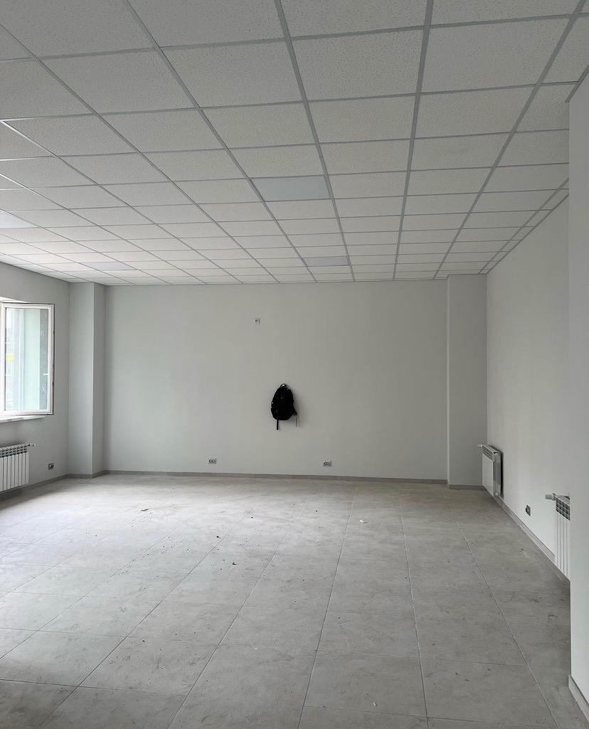 Puste, jasne biuro z szarą podłogą w płytkach, białymi ścianami, podwieszanym sufitem, grzejnikami i plecakiem wiszącym na ścianie.