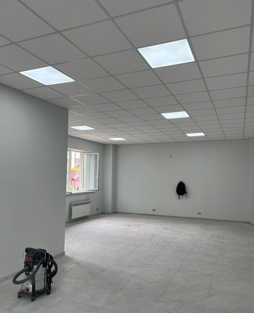 Wnętrze biura po malowaniu ścian na szaro, z zamontowanym sufitem podwieszanym z kwadratowymi panelami LED, widoczny odkurzacz przemysłowy i plecak wiszący na ścianie.