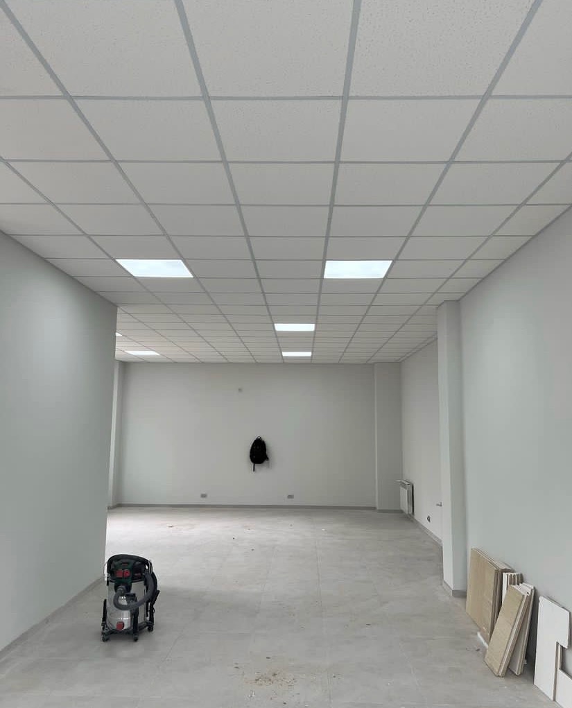 Pusta przestrzeń biurowa z podwieszanym sufitem kasetonowym, widoczne panele podłogowe i odkurzacz przemysłowy, plecak na ścianie.