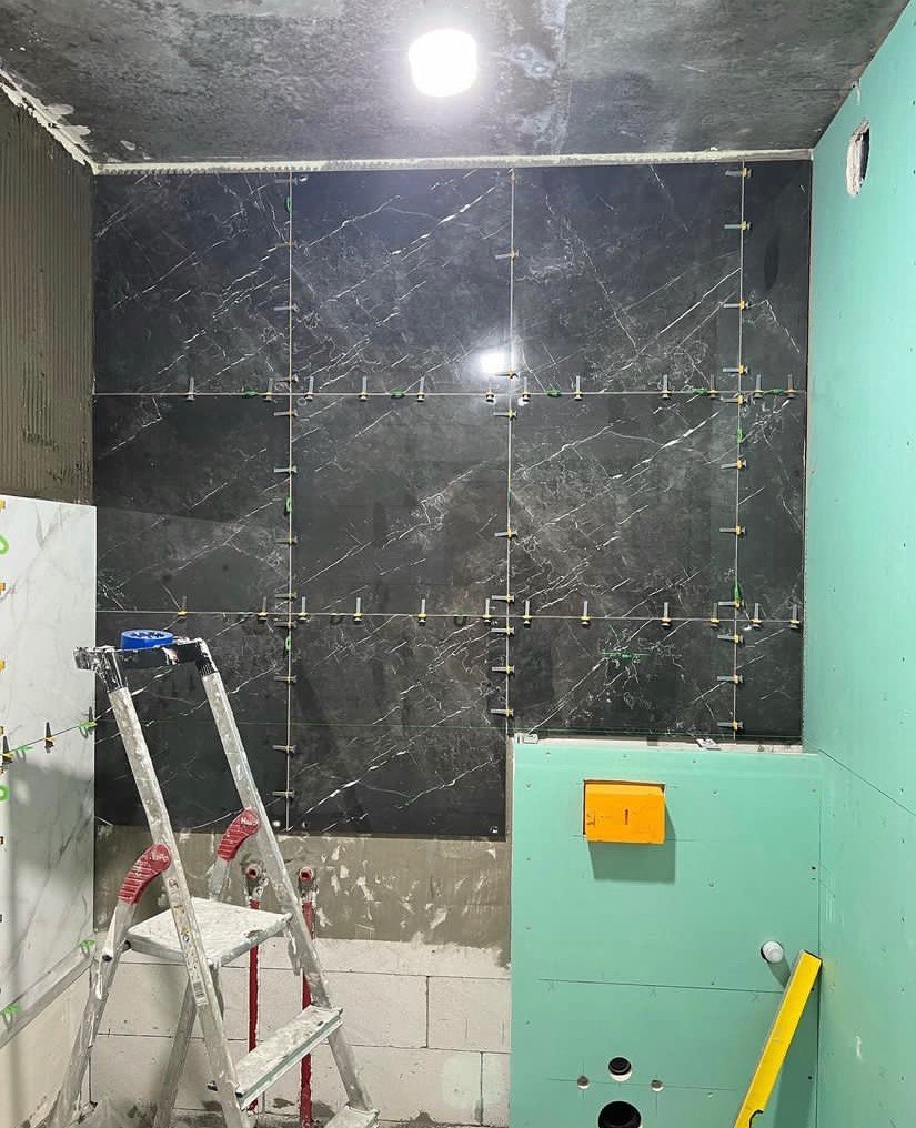 Montaż płytek imitujących czarny marmur na ścianie łazienki, widoczne krzyżyki dystansowe, drabina aluminiowa i niezabudowana ściana z zielonych płyt gipsowo-kartonowych.