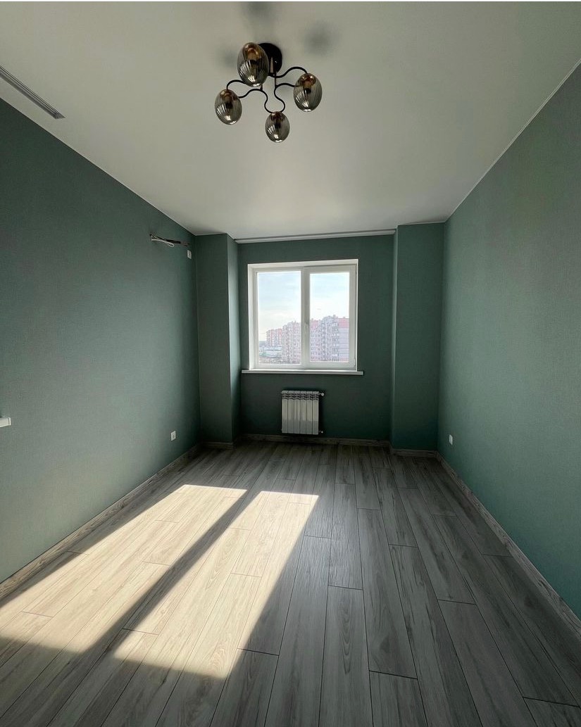 Pomalowane na oliwkowy kolor puste pomieszczenie z panelami podłogowymi, oświetlone promieniami słońca wpadającymi przez okno, z nowoczesną lampą sufitową.