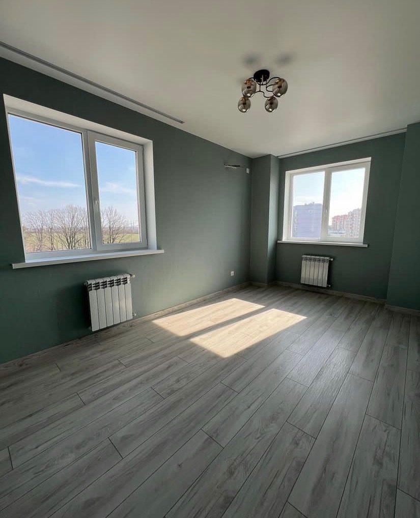 Pusty pokój z pomalowanymi na oliwkowo ścianami, jasnymi panelami podłogowymi i dwoma białymi oknami wpuszczającymi światło słoneczne.
