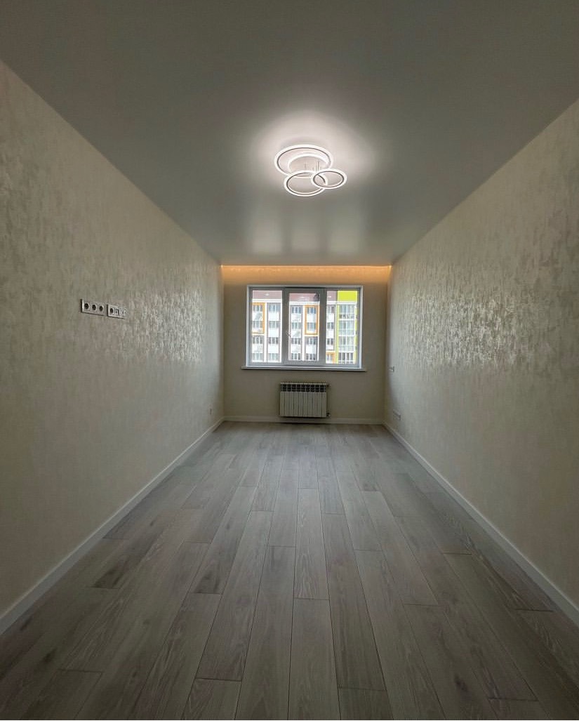Wąskie, puste pomieszczenie po remoncie z panelami podłogowymi w odcieniach szarości, ścianami pokrytymi dekoracyjną fakturą w kolorze kremowym, nowoczesną lampą sufitową i grzejnikiem pod oknem.