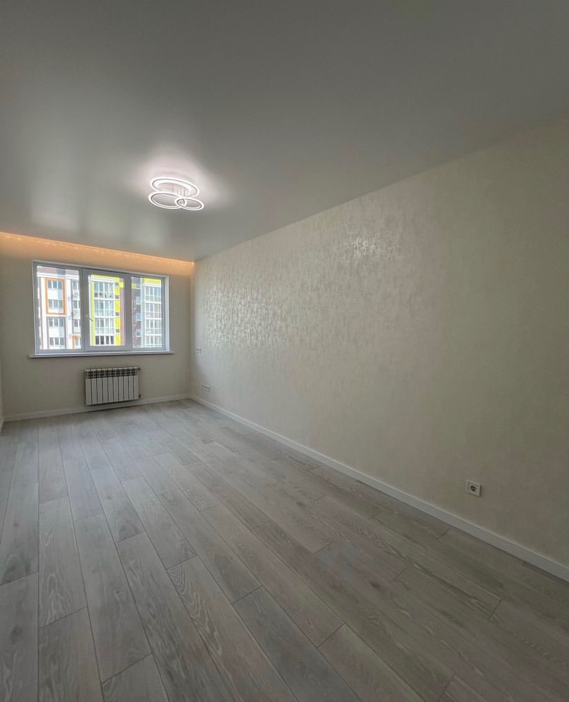 Jasne wnętrze pokoju po remoncie: panele podłogowe w odcieniach szarości, ściany z delikatną fakturą, nowoczesna lampa sufitowa i widok z okna na budynek mieszkalny.