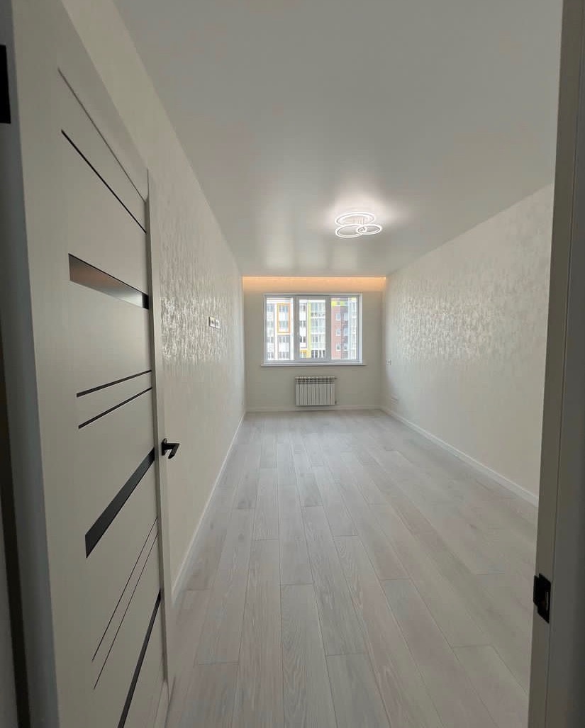 Wnętrze pokoju po remoncie: jasne panele podłogowe, wzorzyste, jasne ściany, białe drzwi z czarnymi wstawkami i nowoczesna lampa sufitowa.