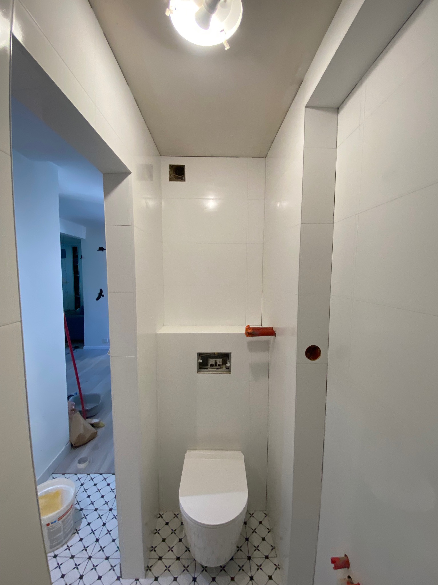 Biała, wąska łazienka w trakcie wykańczania: toaleta, płytki na podłodze i ścianach, widoczne otwory na instalacje. Przez otwarte drzwi widać fragment mieszkania.