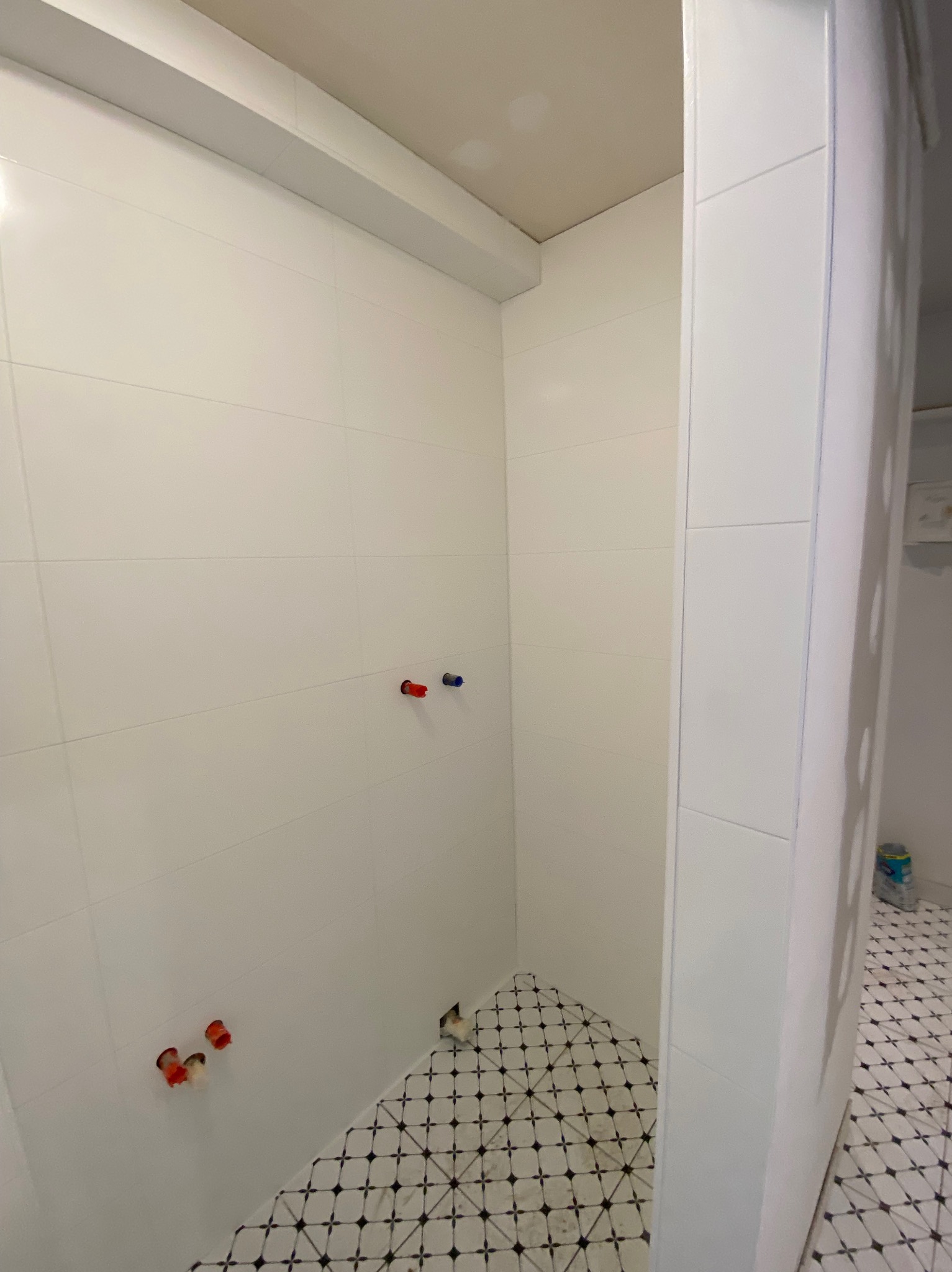 Biała łazienka z ułożonymi płytkami podłogowymi w czarno-białą kratkę. Widoczne przyłącza wodne na ścianie i białe płytki ścienne. Minimalistyczny design.