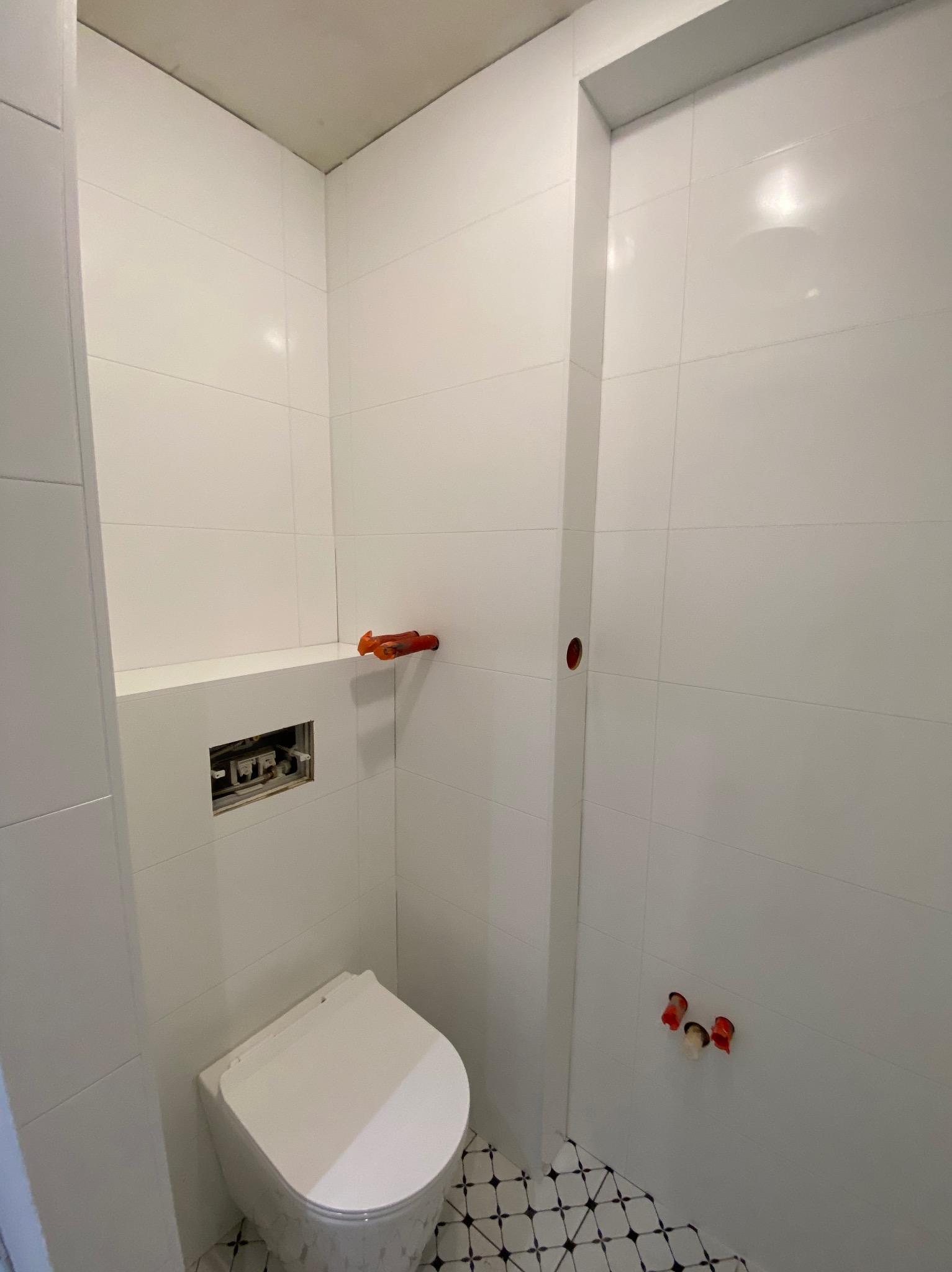 Biała, wykończona łazienka z wiszącą toaletą i wzorzystą podłogą. Widoczne przyłącza wodne i otwór na stelaż podtynkowy. Minimalistyczny design.