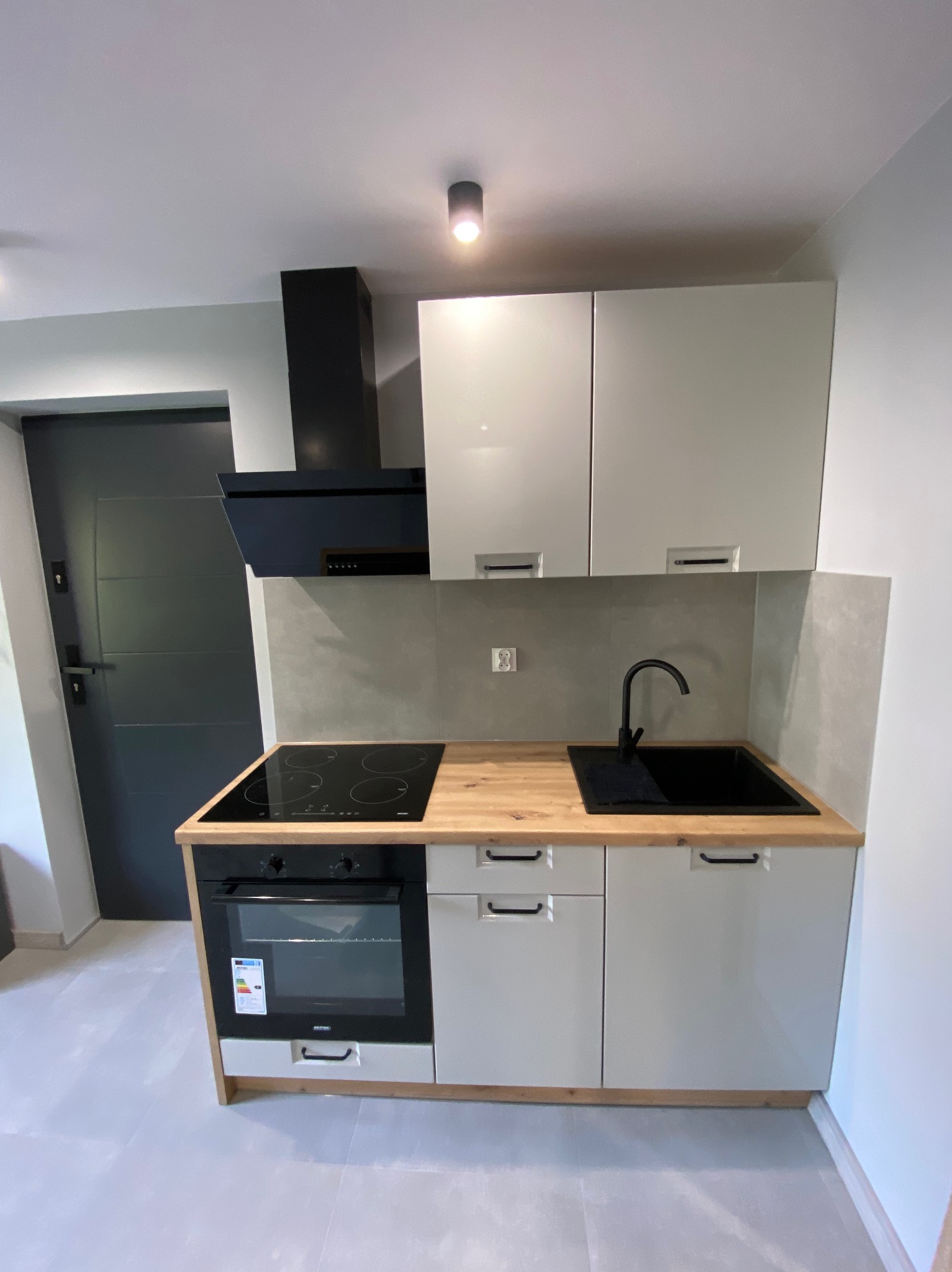 Nowoczesna, jasna kuchnia z drewnianym blatem, czarnym zlewem i płytą indukcyjną. Minimalistyczny design, szare ściany i podłoga. Czarny okap nad płytą.