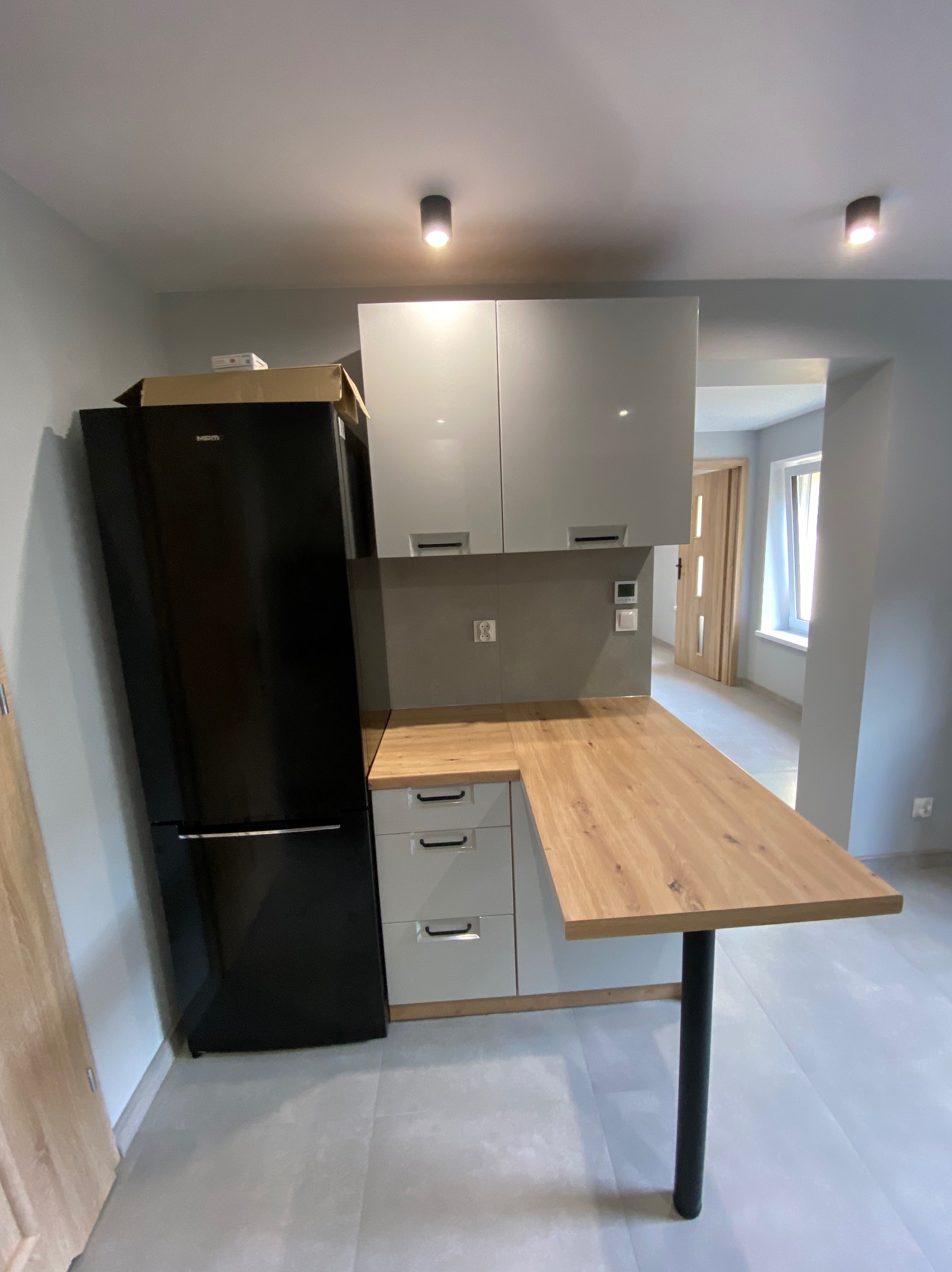 Nowoczesna kuchnia z czarną lodówką, białymi szafkami i drewnianym blatem, doświetlona punktowymi lampami sufitowymi. Widok na dalszą część mieszkania.