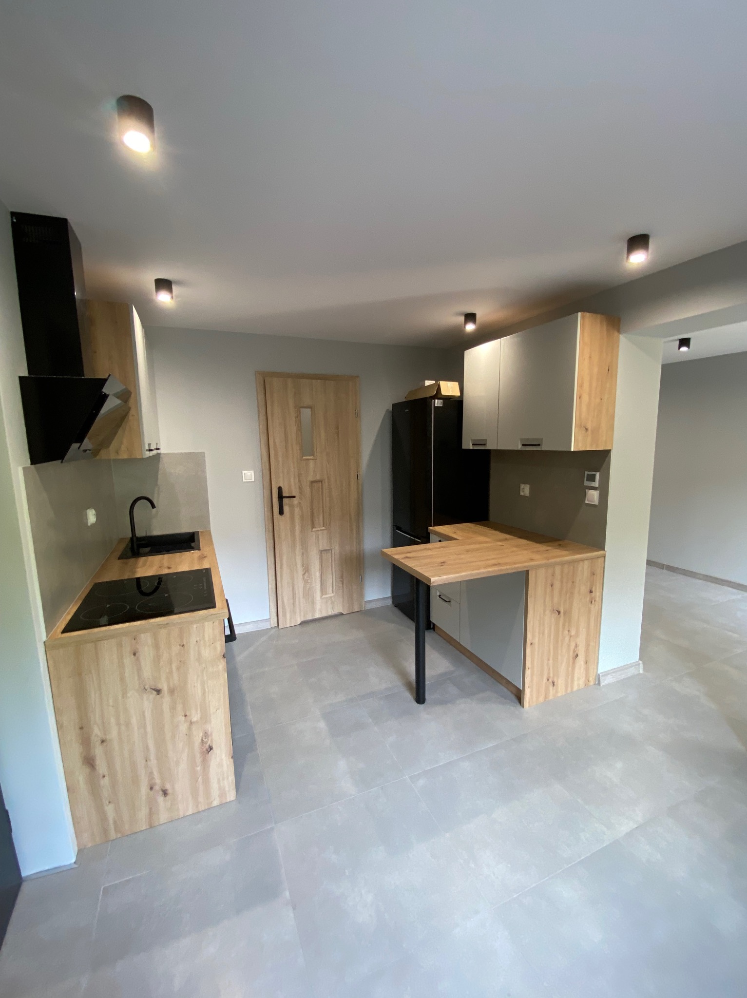 Nowoczesna kuchnia z drewnianymi akcentami: zabudowa kuchenna z blatem, czarna płyta indukcyjna i okap, szare płytki podłogowe, minimalistyczne oświetlenie.