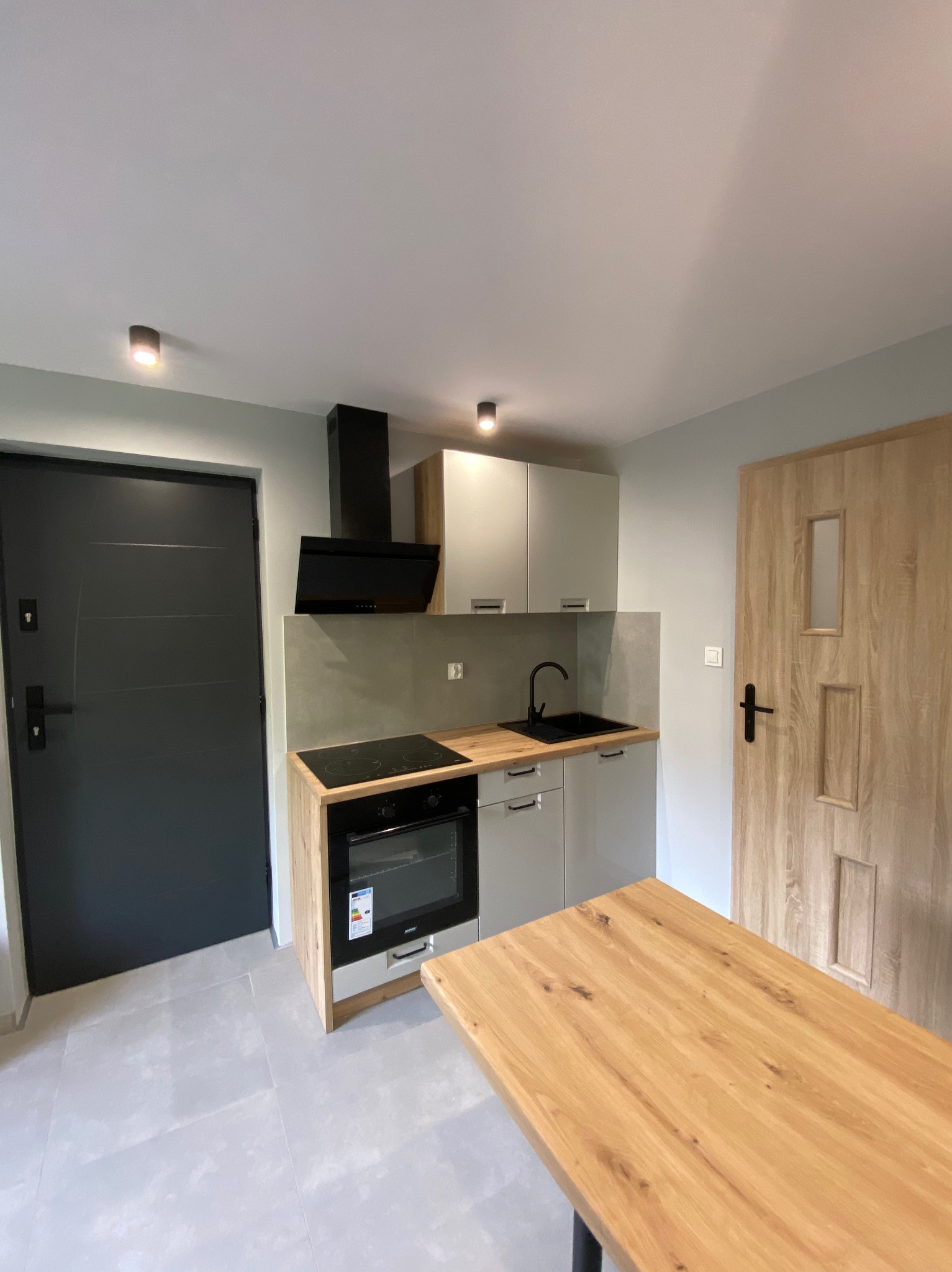 Jasna kuchnia po remoncie: szare szafki, drewniany blat, czarny okap i płyta indukcyjna. Na podłodze szare płytki, widoczne drzwi wejściowe i wewnętrzne.