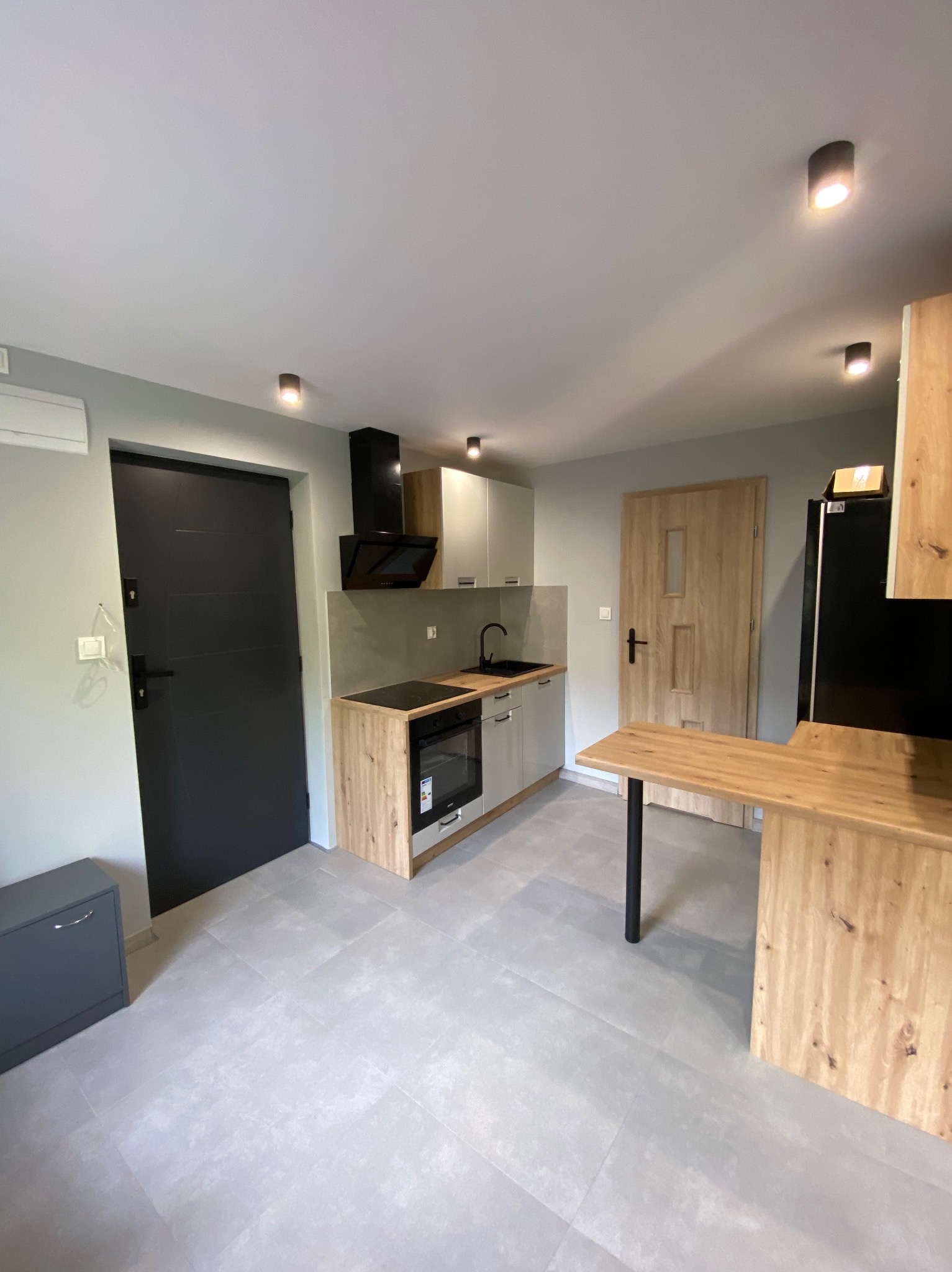 Jasna kuchnia z drewnianymi akcentami: zabudowa kuchenna, wyspa z blatem, czarne AGD i szara podłoga z płytek. Nowoczesne oświetlenie punktowe.