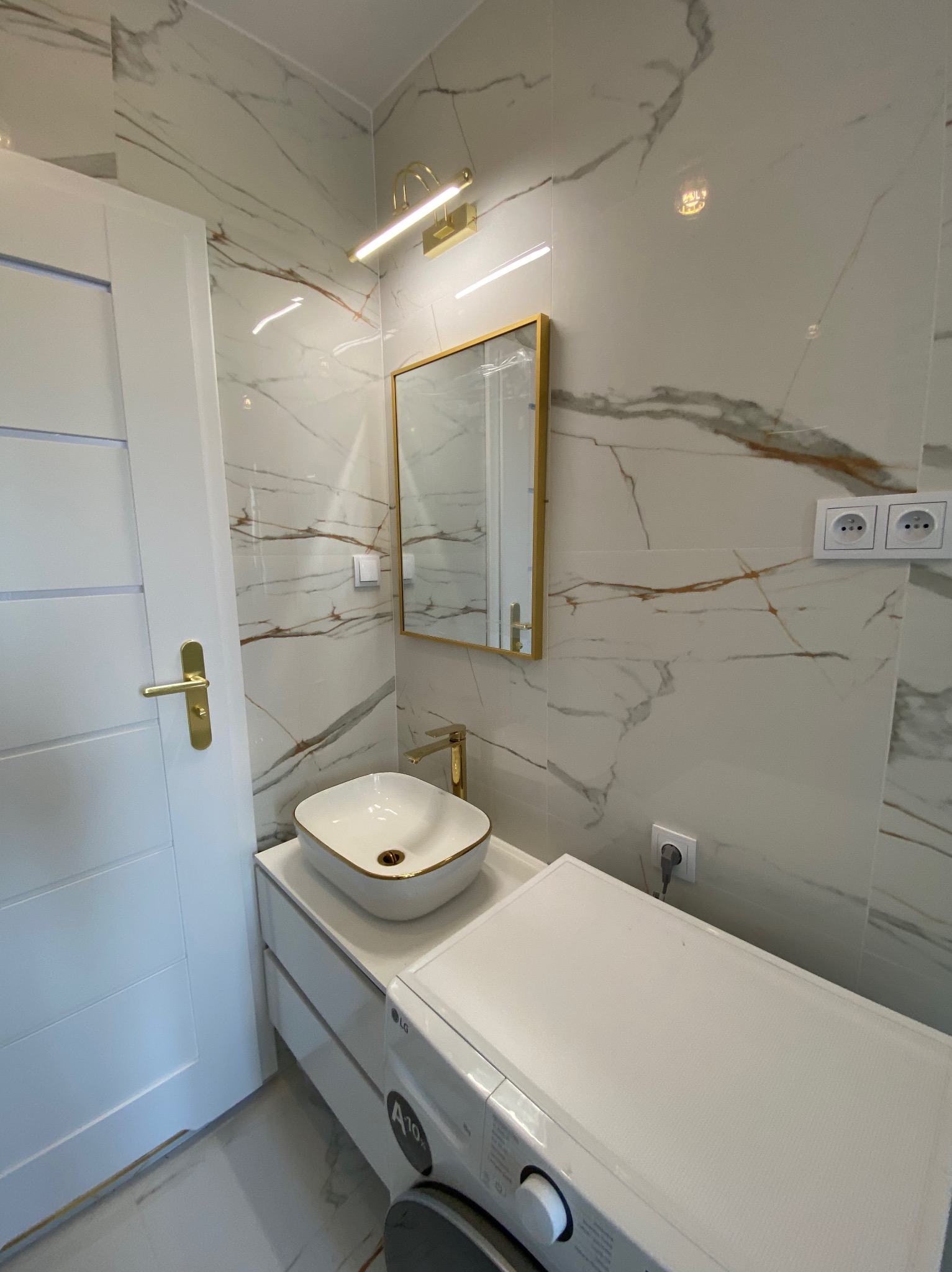 Biała łazienka z marmurowymi płytkami, złotymi akcentami i nowoczesną umywalką nablatową. Widoczna pralka pod blatem. Oświetlenie LED nad lustrem.