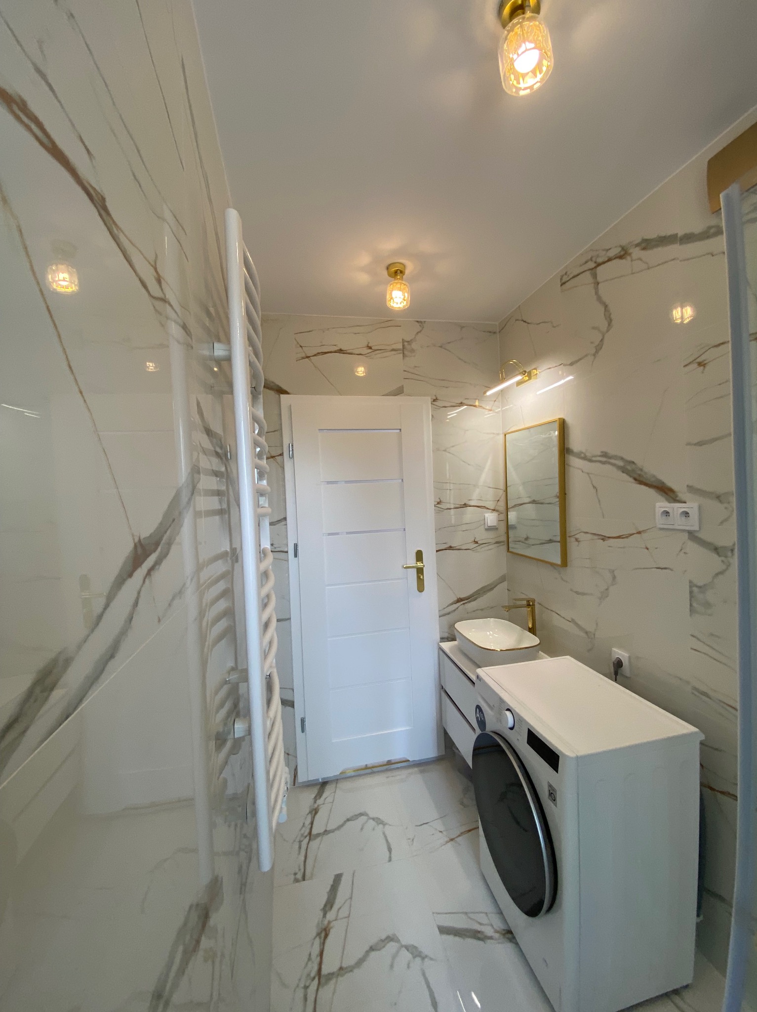 Wykończona łazienka w Opolu z marmurowymi płytkami, złotymi akcentami i nowoczesną pralką. Widoczny grzejnik drabinkowy i lustro z oświetleniem.