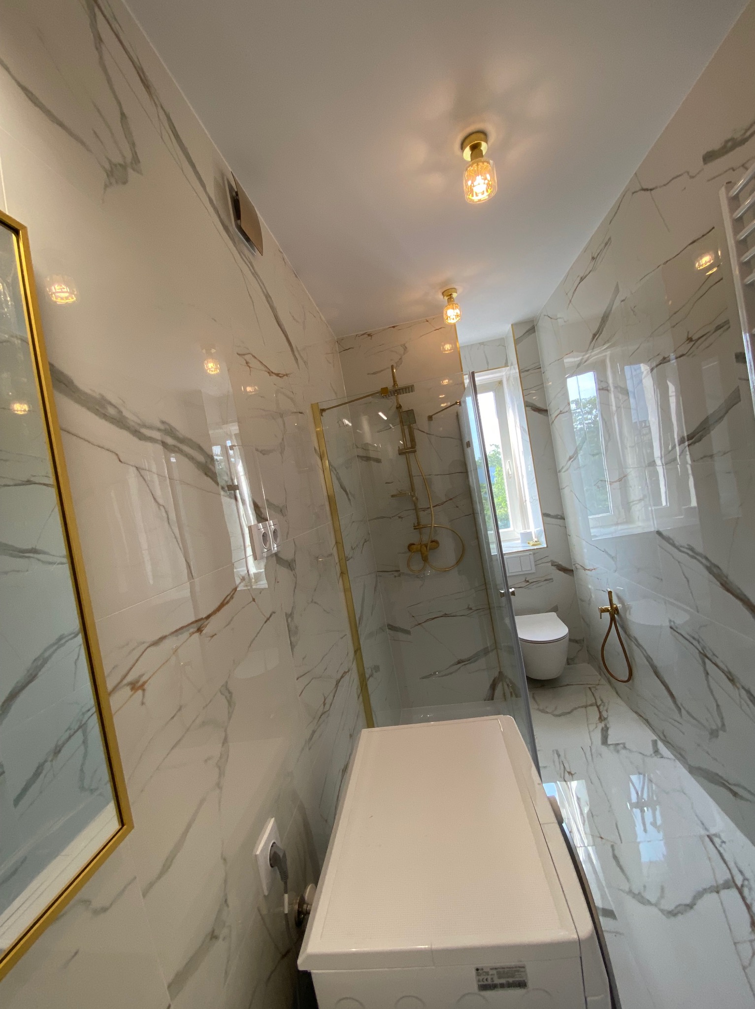 Wykończona łazienka z kabiną prysznicową w złotej ramie, marmurowe płytki na ścianach i podłodze, widoczna pralka i toaleta. Oświetlenie sufitowe.