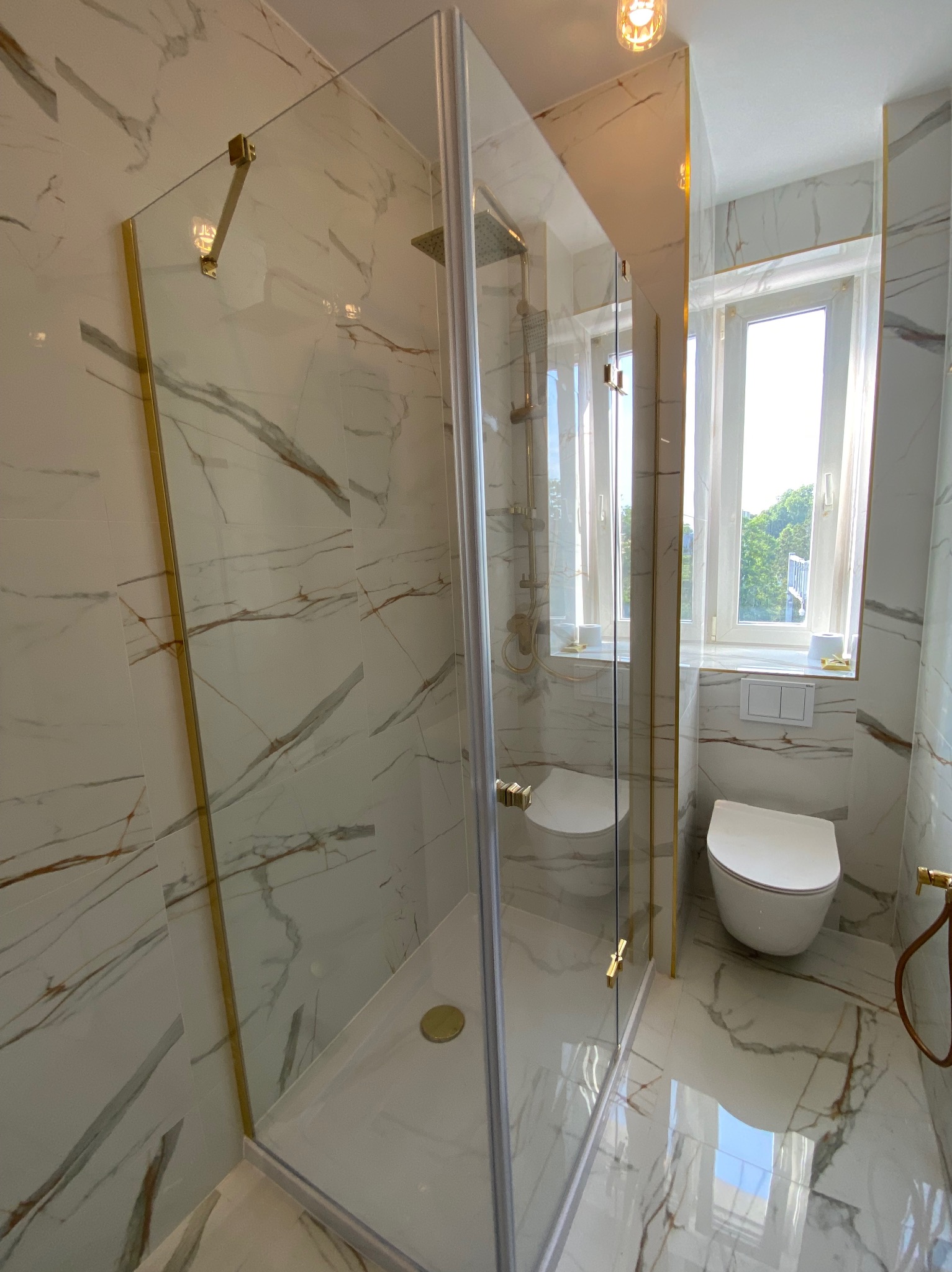 Nowoczesna łazienka z kabiną prysznicową typu walk-in, wykończona białym marmurem z złotymi akcentami. Widoczne WC i okno z widokiem na zieleń.