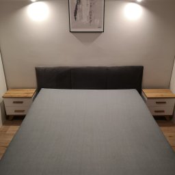 Piotr Szczepanek - Nowoczesna sypialnia z szarym łóżkiem, drewnianymi szafkami nocnymi i abstrakcyjnym obrazem na ścianie, oświetlona dwoma punktowymi lampami.