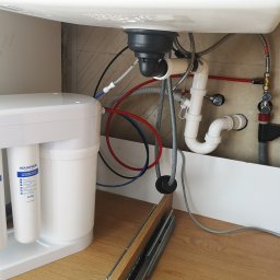Piotr Szczepanek - Montaż systemu filtracji wody Aquaphor pod zlewem kuchennym, widoczne rury doprowadzające i odprowadzające wodę, oraz wkłady filtrujące K2 i K7M.