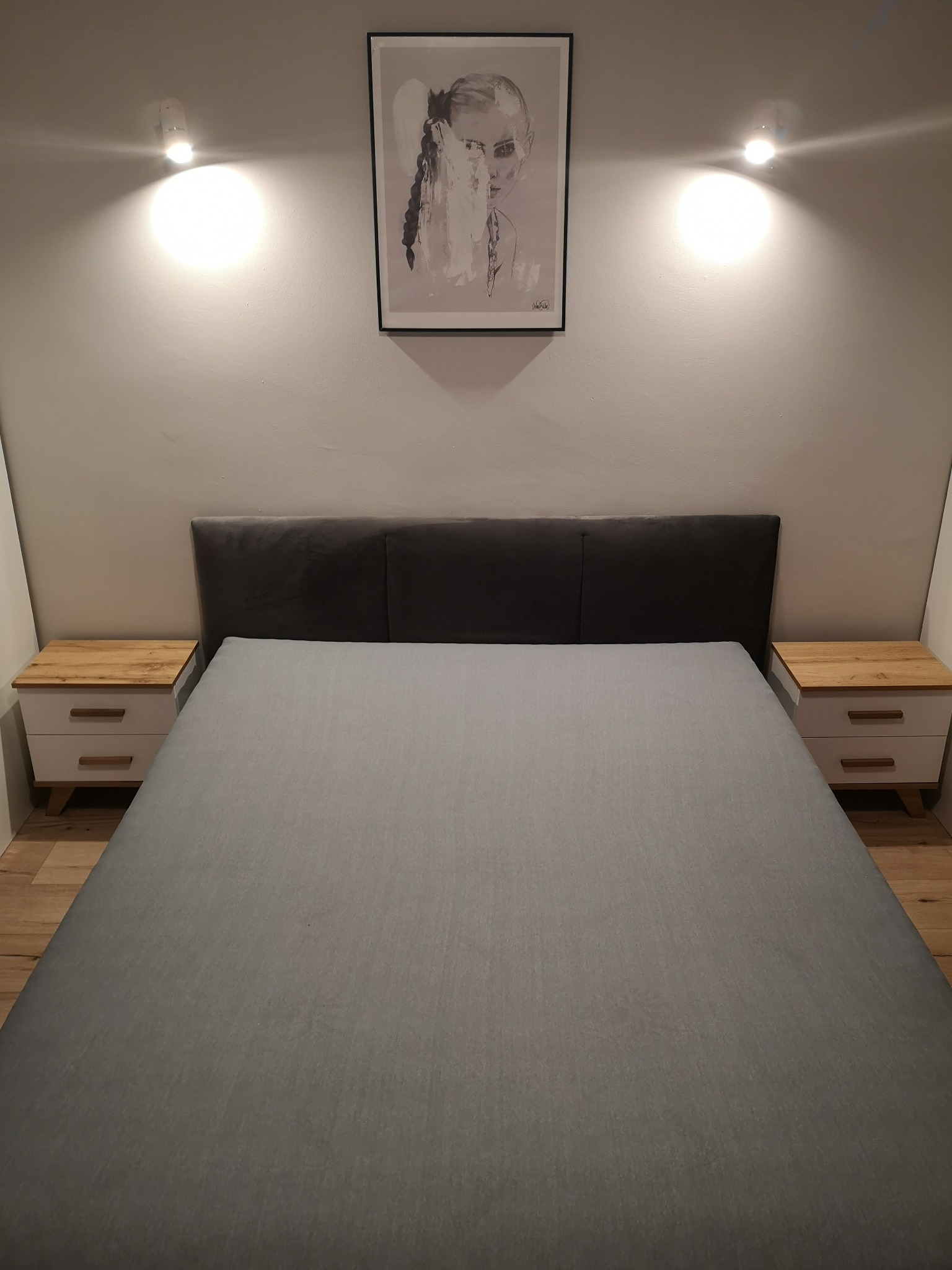 Nowoczesna sypialnia z szarym łóżkiem, drewnianymi szafkami nocnymi i abstrakcyjnym obrazem na ścianie, oświetlona dwoma punktowymi lampami.