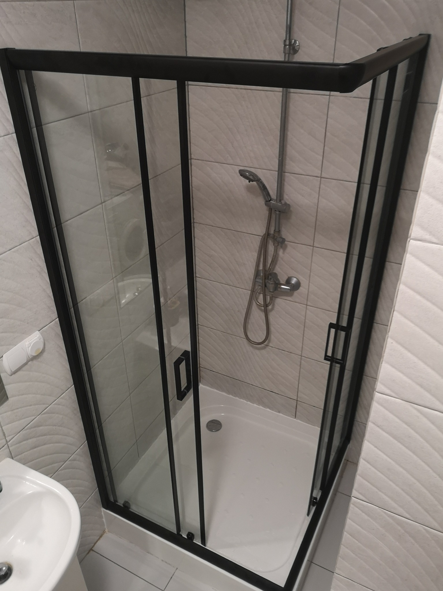 Nowoczesna kabina prysznicowa z czarnymi ramami i brodzikiem, widoczna armatura i umywalka w tle, płytki imitujące fale na ścianach.