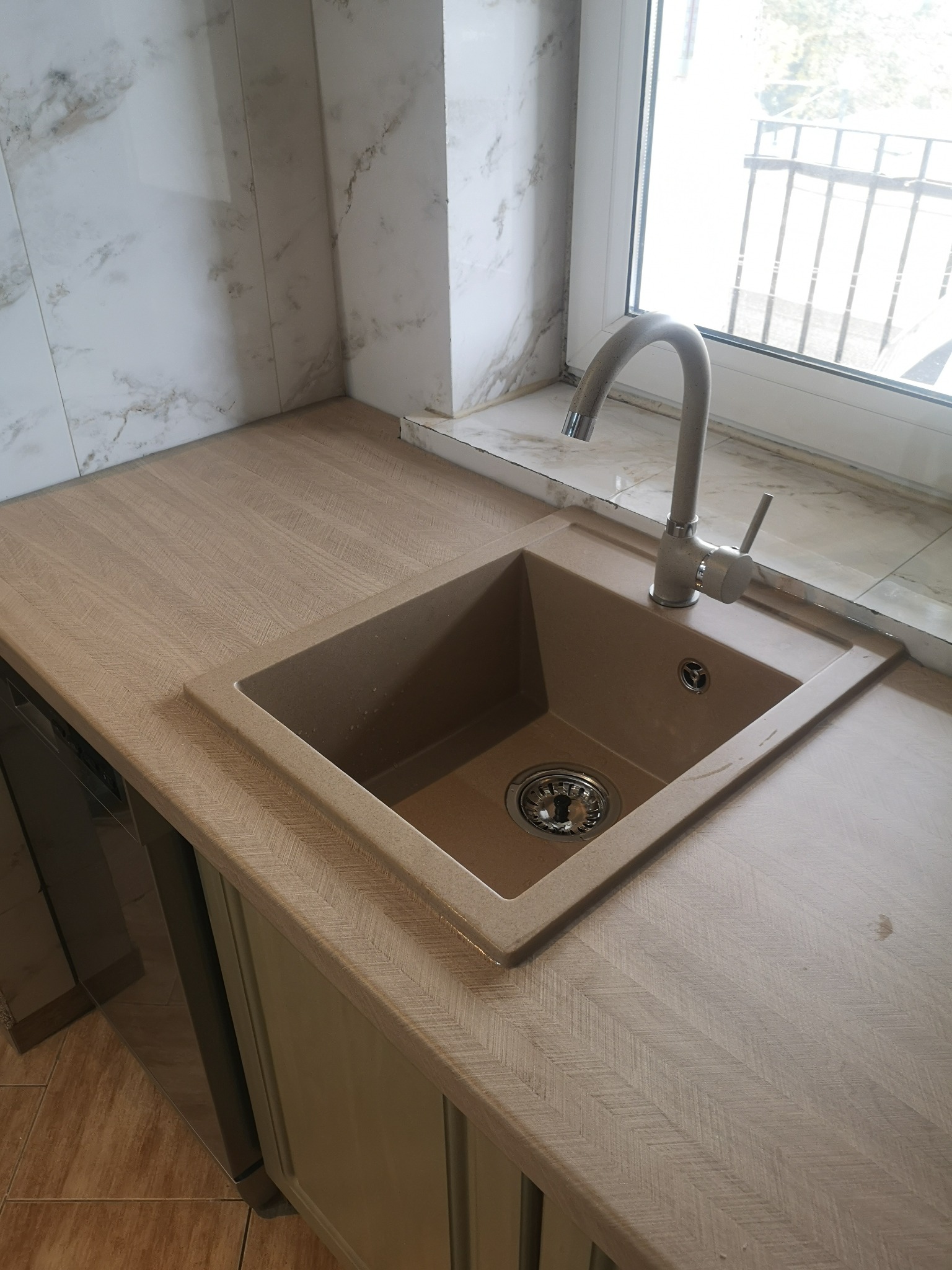 Beżowy zlew granitowy z chromowaną baterią wkomponowany w blat o strukturze drewna, w kuchni po remoncie w Ostrołęce, widok z góry, naturalne światło.