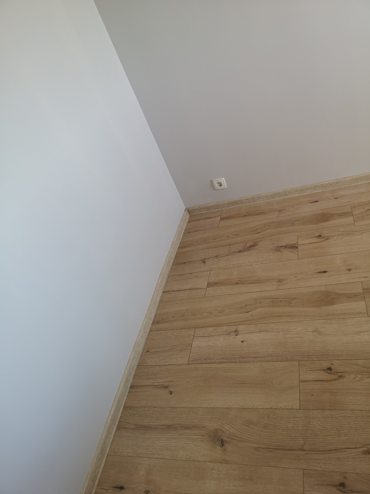 Jasne panele podłogowe ułożone wzdłuż białej ściany z zamontowanym gniazdkiem elektrycznym przy listwie przypodłogowej.