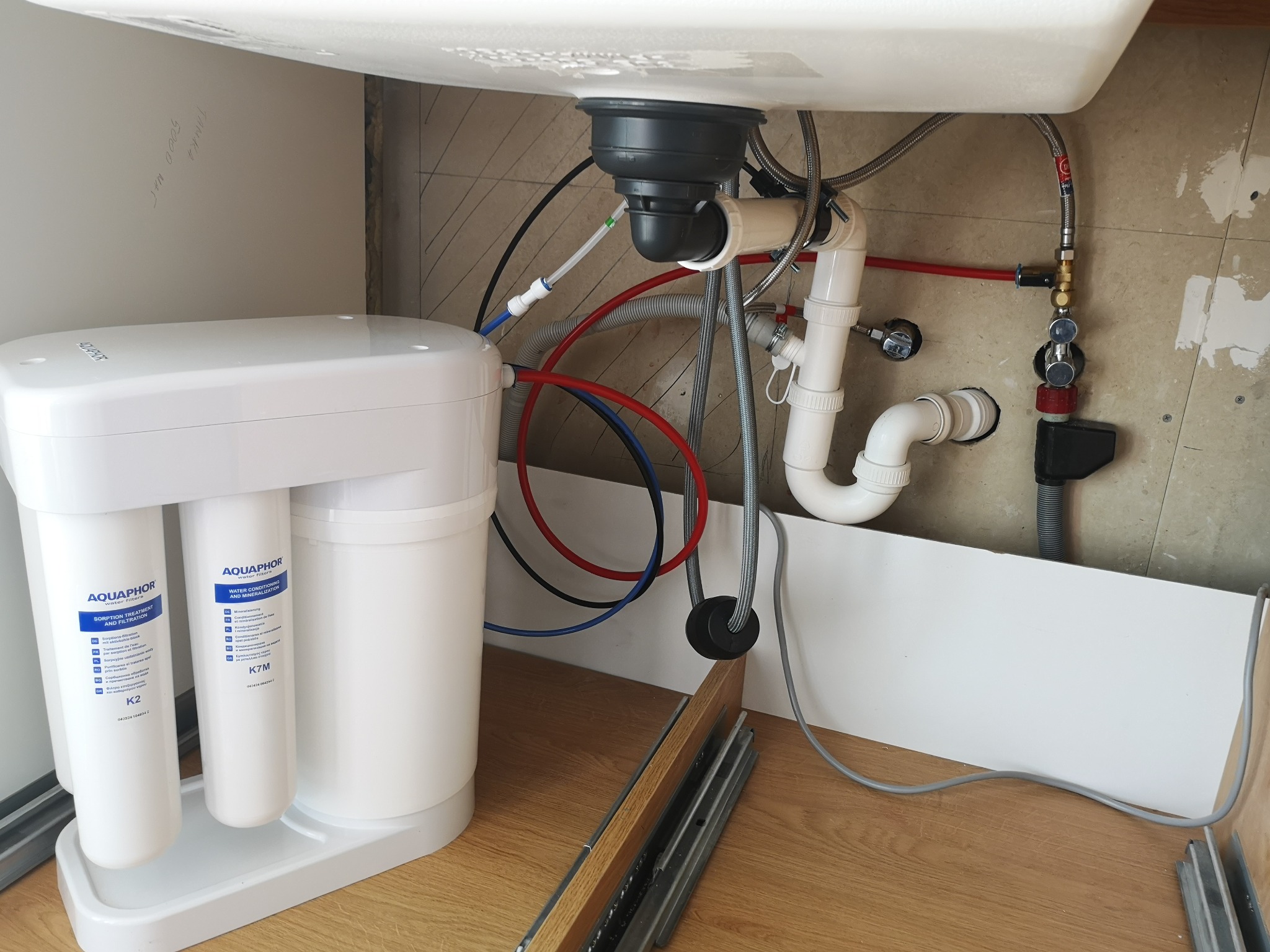 Montaż systemu filtracji wody Aquaphor pod zlewem kuchennym, widoczne rury doprowadzające i odprowadzające wodę, oraz wkłady filtrujące K2 i K7M.