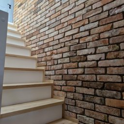 Firma Transportowo-Budowlana Tomasz Kostrzewa - Schody z drewnianymi stopniami i białą podstopnicą, przylegające do ściany z dekoracyjnej cegły w odcieniach brązu i beżu, oświetlone naturalnym światłem.