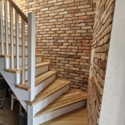 Firma Transportowo-Budowlana Tomasz Kostrzewa - Drewniane schody z białymi balustradami, obok ściana z cegły w odcieniach brązu i beżu. Widok z dołu, ukazujący zakręt schodów i fakturę ceglanej ściany.