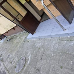 Firma Transportowo-Budowlana Tomasz Kostrzewa - Wejście do budynku z brązowymi drzwiami i szarymi płytkami na schodach oraz chodniku. Widoczna metalowa poręcz i fragment elewacji. Ujęcie z dołu, obrócone.