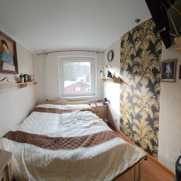 Firma Transportowo-Budowlana Tomasz Kostrzewa - Przytulna sypialnia z łóżkiem, wzorzystą tapetą w palmy i drewnianymi meblami. Na ścianach obrazy i półki z dekoracjami. Widok z okna na zewnątrz.