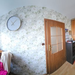 Firma Transportowo-Budowlana Tomasz Kostrzewa - Pokój po remoncie: ściana w kwiaty, drewniane drzwi z szybą, duży zegar na ścianie, szafa przesuwna i komoda. Widok na okno z parapetem i kwiatami.