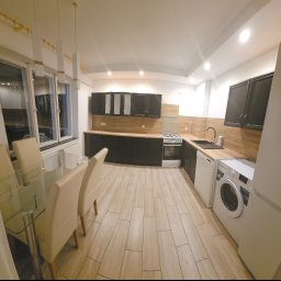 Firma Transportowo-Budowlana Tomasz Kostrzewa - Nowoczesna kuchnia z czarnymi szafkami i drewnianym blatem w Czarnkowie. Widoczny stół z krzesłami, sprzęt AGD i jasna podłoga.