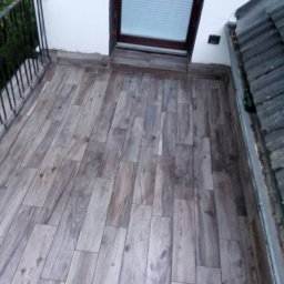 Firma Transportowo-Budowlana Tomasz Kostrzewa - Wykończony balkon z drewnopodobną podłogą z płytek, czarną balustradą i oknem dachowym. Widok z góry, ukazujący detale ułożenia płytek i estetykę wykończenia.