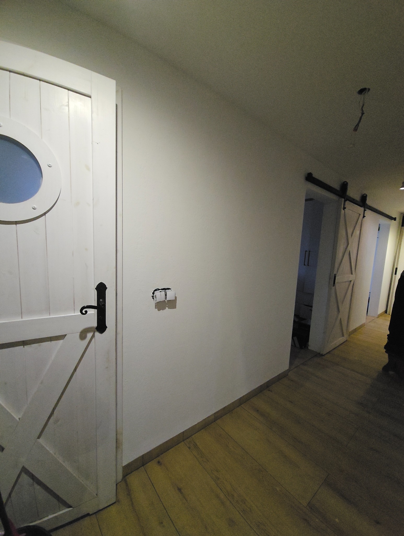 Białe, drewniane drzwi z okrągłym okienkiem i czarną klamką w jasnym korytarzu. W tle widoczne przesuwne drzwi stodołowe i panele podłogowe.