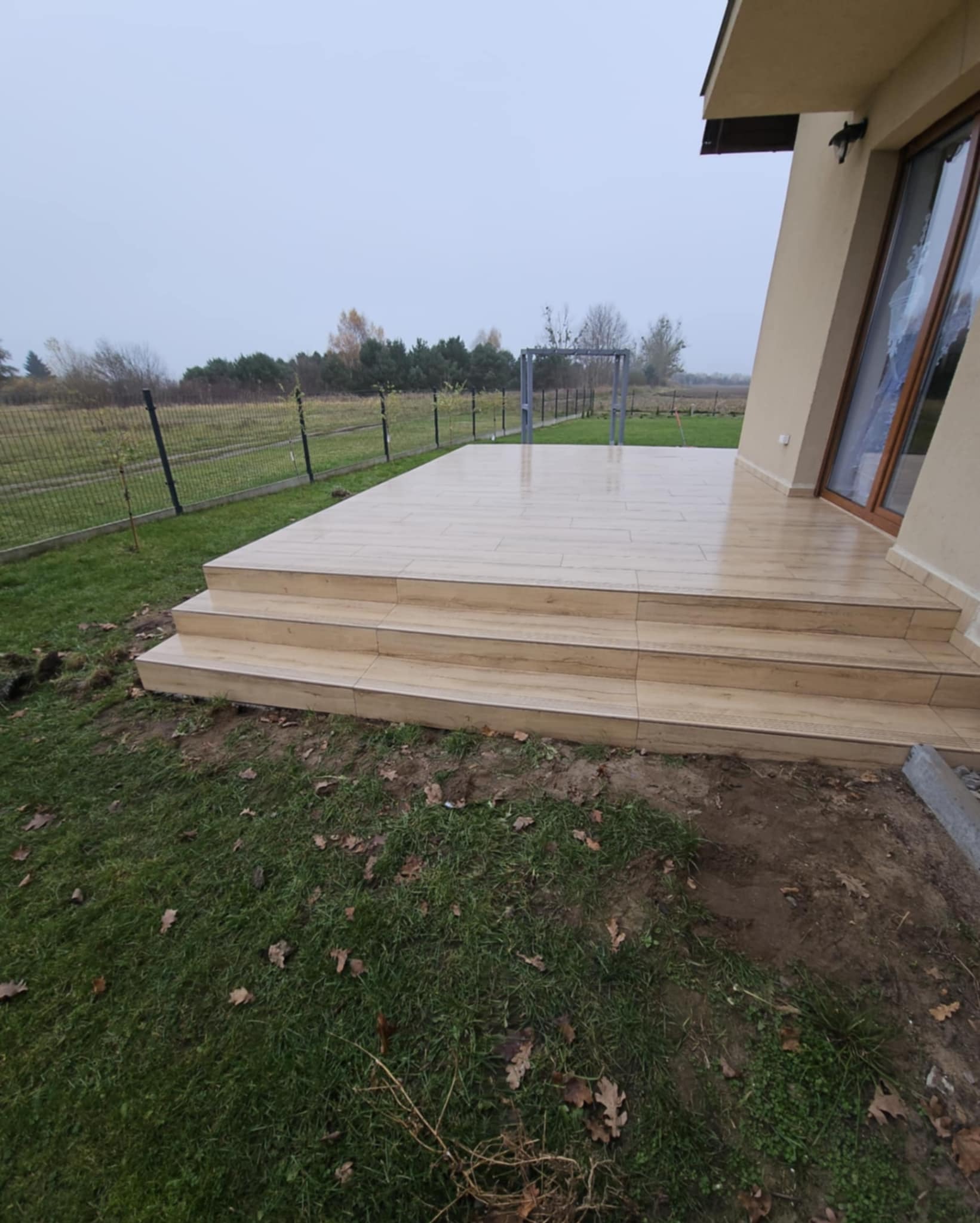 Wyłożony płytkami taras z trzema schodkami, przylegający do budynku z oknem. Widok na zewnątrz, na trawnik i drzewa w tle. Płytki w odcieniach beżu z delikatnym wzorem drewna.