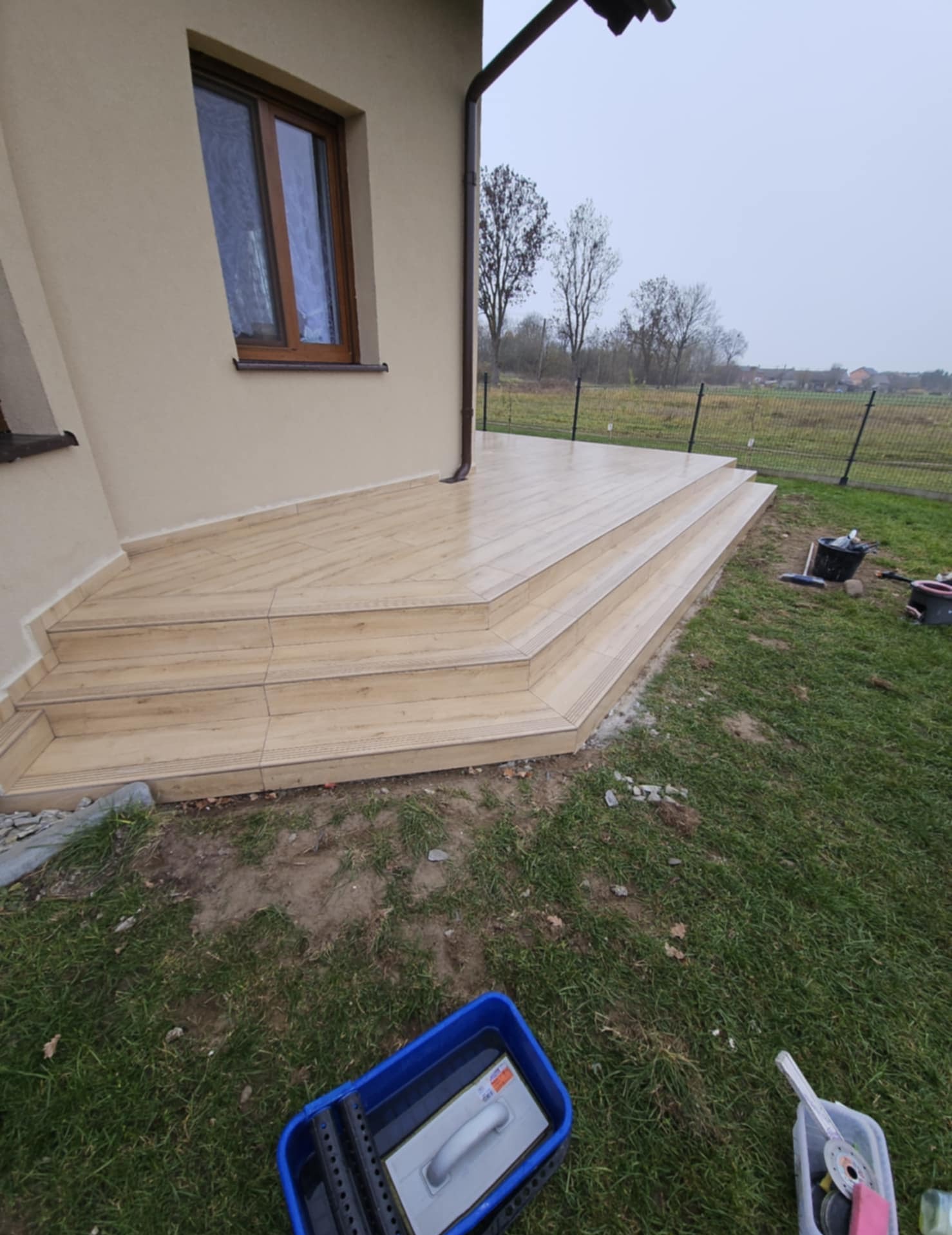 Wykończony taras z beżowymi płytkami imitującymi drewno, prowadzący do domu z brązowymi oknami. Widoczne narzędzia glazurnicze i trawa.