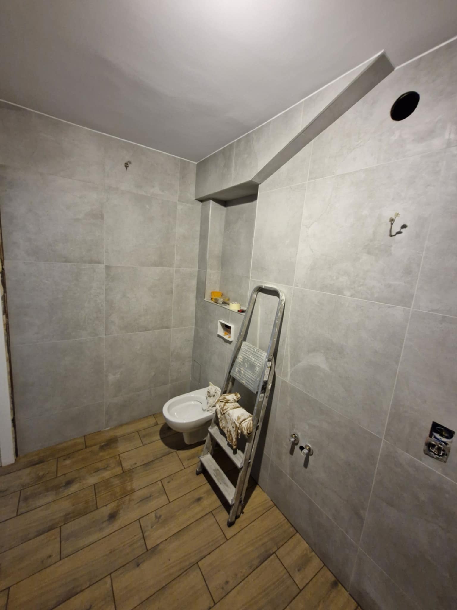 Szara łazienka w trakcie remontu: ułożone płytki ścienne i podłogowe imitujące drewno, drabina oparta o ścianę, częściowo zamontowana toaleta. Widoczne niedokończone detale.