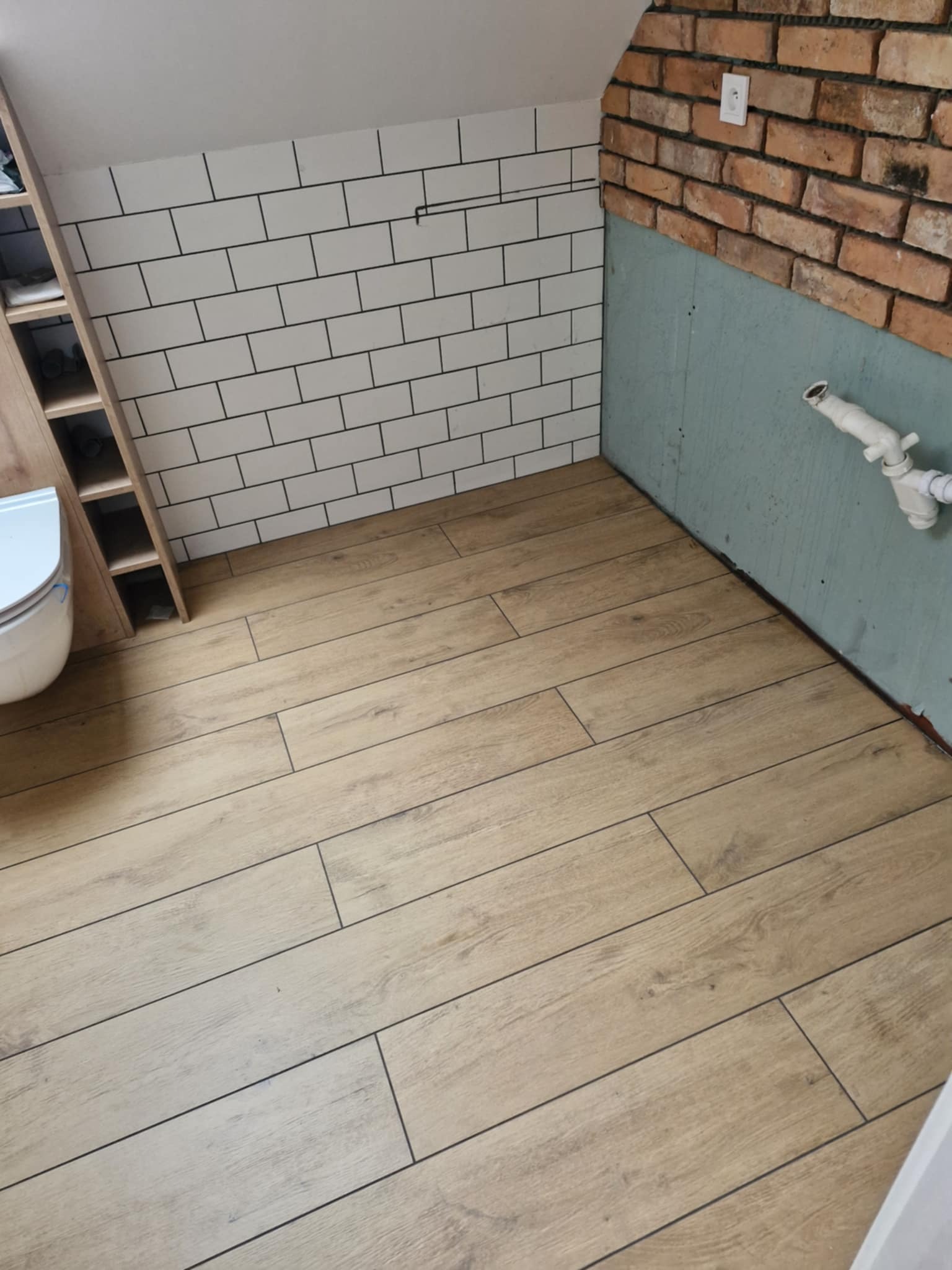 Ukończona łazienka w trakcie remontu: jasne panele podłogowe imitujące drewno, białe płytki ścienne typu metro z czarną fugą i fragment ściany z cegły.